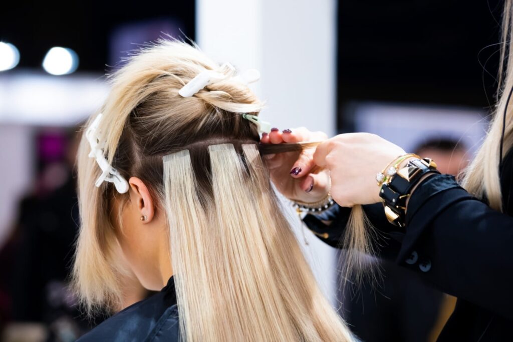 A hairstylist at a hair studio in Winter Haven is applying hair extensions to a woman's head using clips and tape, with sectioned hair held in place by white hair clips.