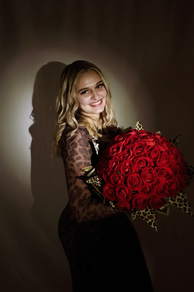 A woman with long blonde hair, styled at a hair studio in Winter Haven, wears a sheer animal print top and smiles while holding a large bouquet of red roses against a neutral background.