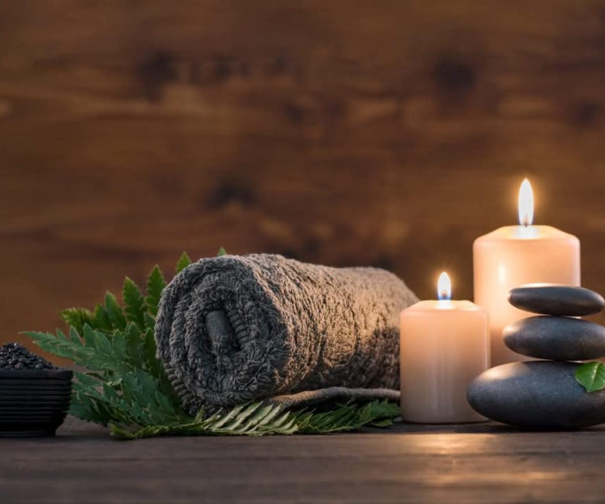 A rolled towel, two lit candles, stacked smooth stones, a small bowl of black pebbles, and green fern leaves create a calming ViBE on a wooden surface—perfect for your Winter Haven retreat.