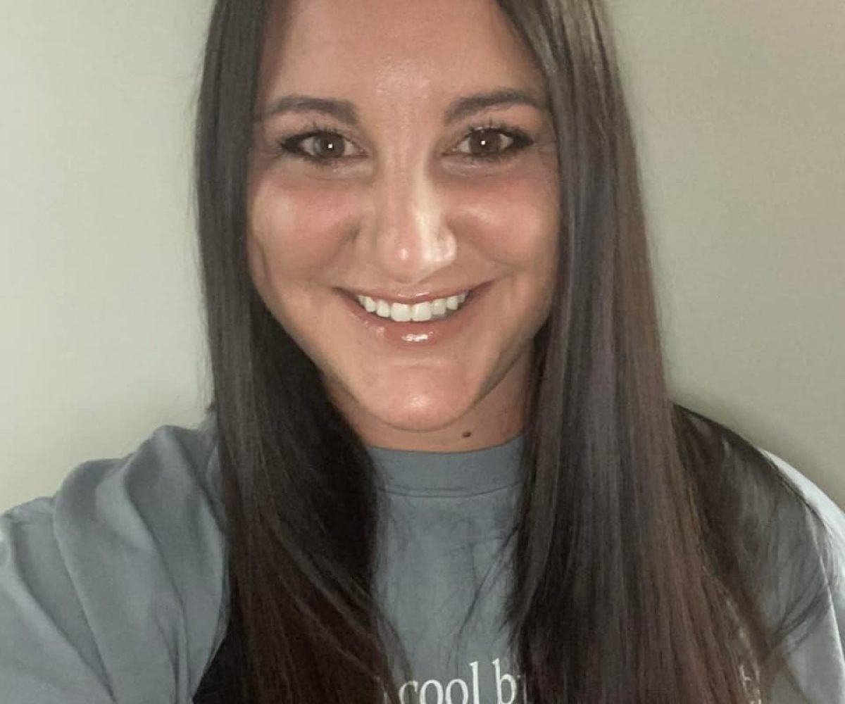 A woman with long straight brown hair and a grey shirt smiles at the camera in front of a plain light-colored background, radiating a friendly ViBE reminiscent of Winter Haven warmth.