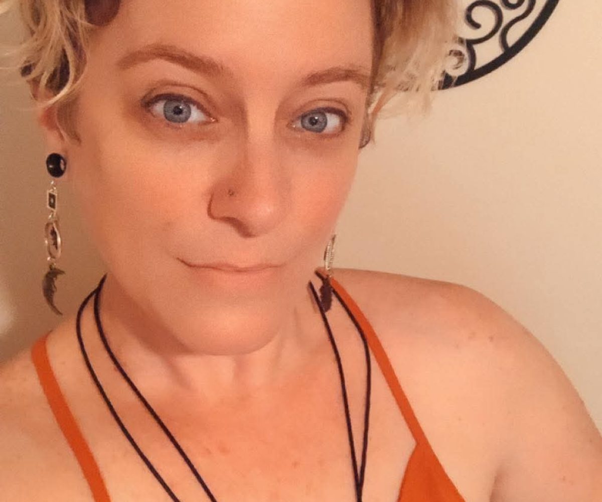 A person with short blond hair wearing an orange sleeveless top and layered necklaces poses indoors in front of a decorative wall piece, radiating ViBE style in true Winter Haven fashion.