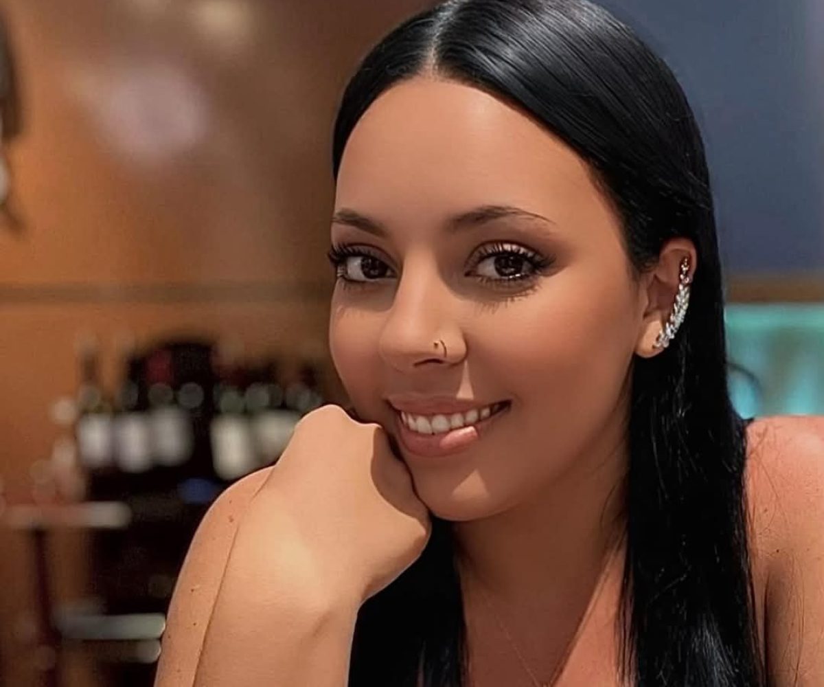 A woman with long black hair, wearing a nose ring and ear cuff, smiles at the camera in a dimly lit Winter Haven setting with wine bottles in the background, capturing the chic ViBE of the moment.