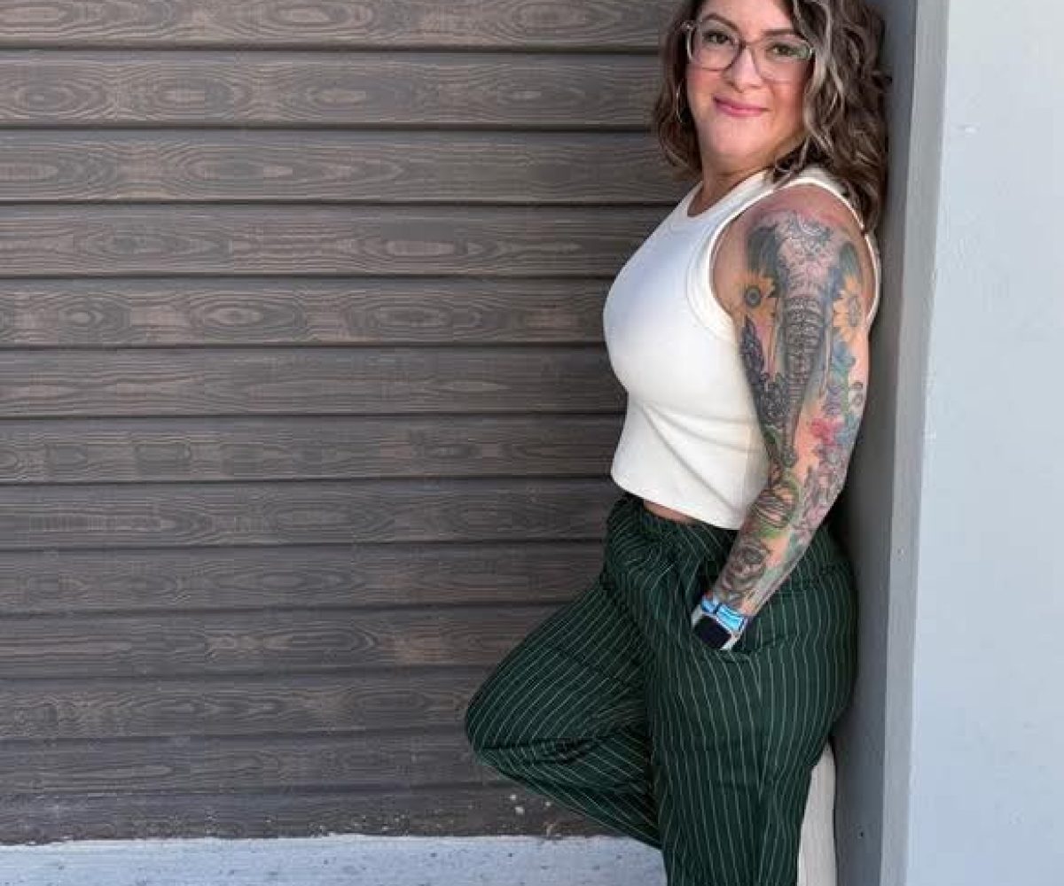 A person with tattoos on their left arm leans against a wall in a ViBE-inspired look, wearing a white sleeveless top, green pinstriped pants, and white shoes, set against a neutral background reminiscent of Winter Haven cool.